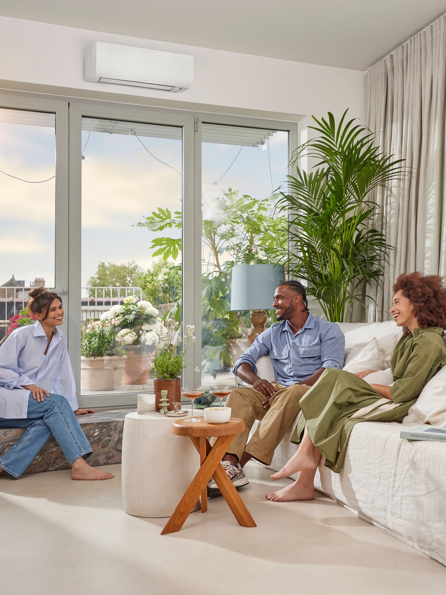 En man och två kvinnor sitter och samtalar i ett ljust och luftigt rum med vita väggar och en diskret, vit väggmonterad luft/luftvärmepump ovanför ett stort panoramafönster en solig dag