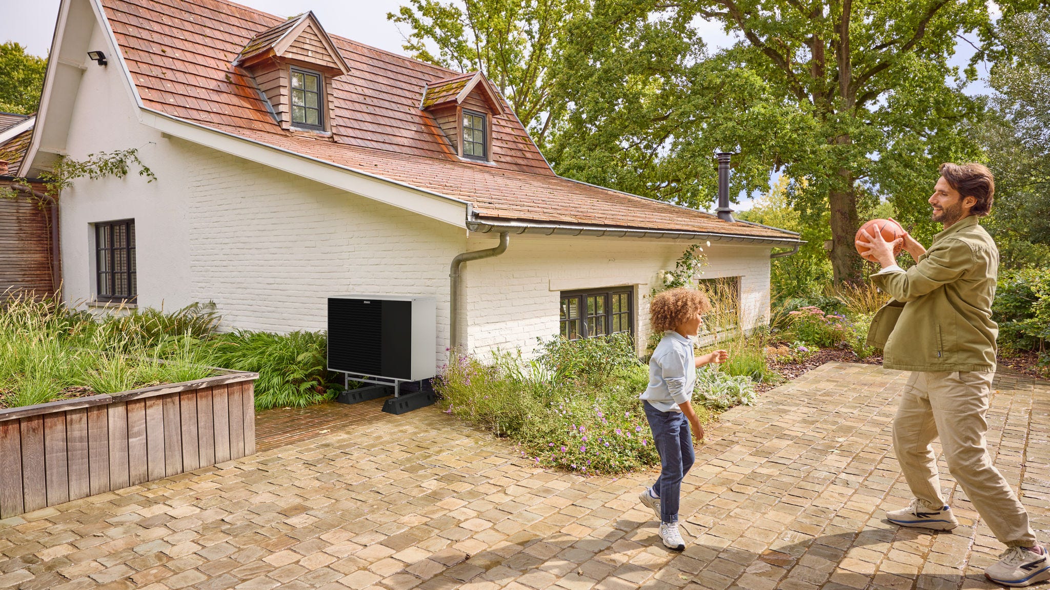 En pappa som leker boll med sitt barn ute på gården, med en värmepump installerad i närheten