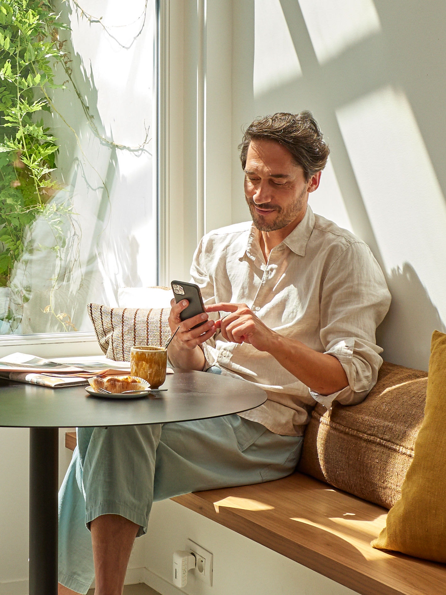 En man som kopplar av och tittar på sin telefon sittandes vid ett bord med en kopp kaffe och lite mat framför sig