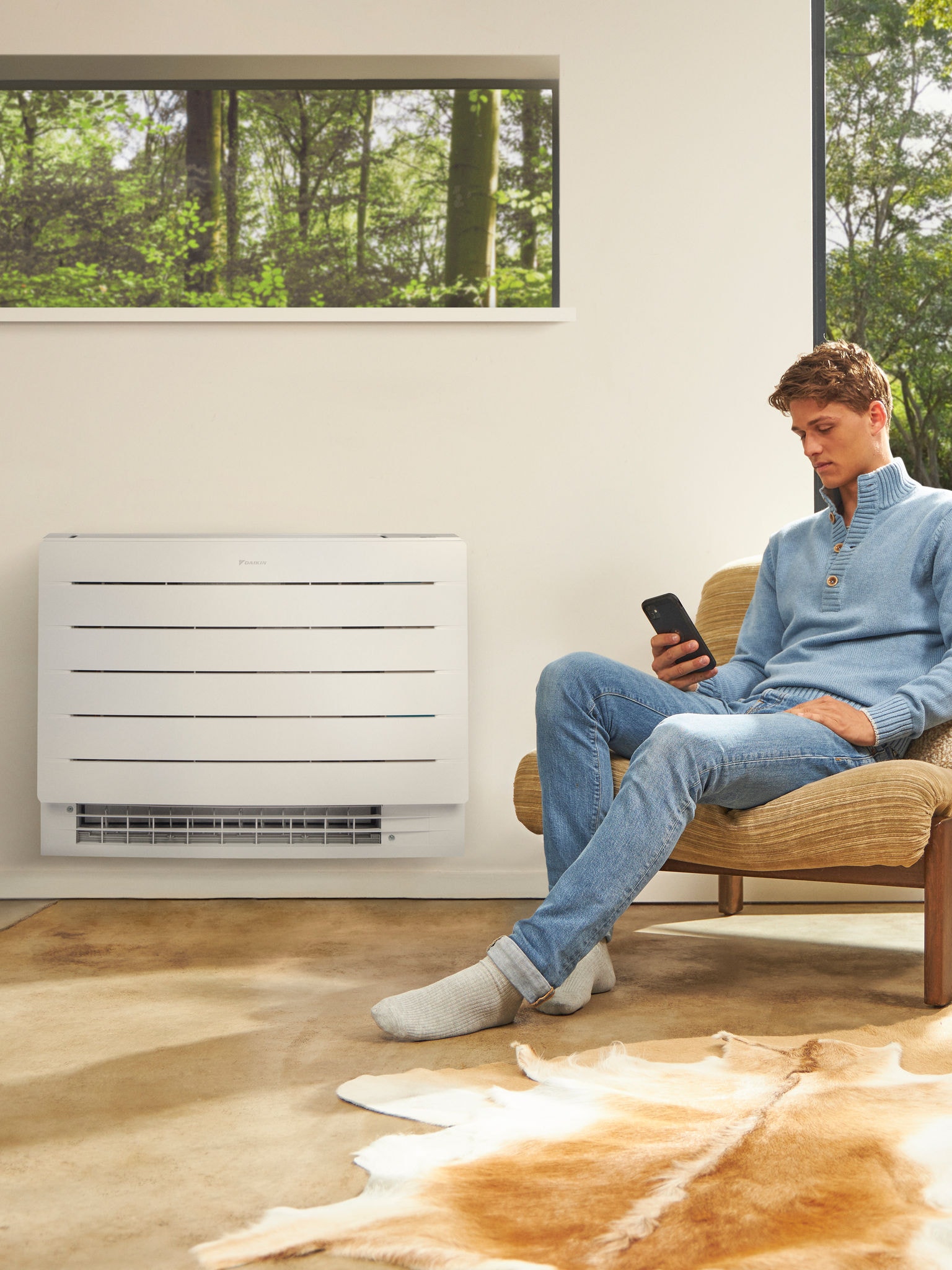 En man sitter i ett ljust rum och tittar på sin telefon, med en luft/luftvärmepump monterad på väggen bakom honom