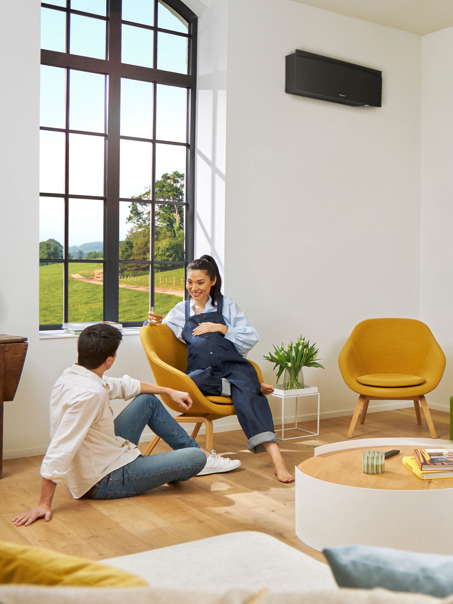 En man och en kvinna sitter framför ett stort fönster i ett ljust rum, med en svart väggmonterad värmepump på väggen bakom dem