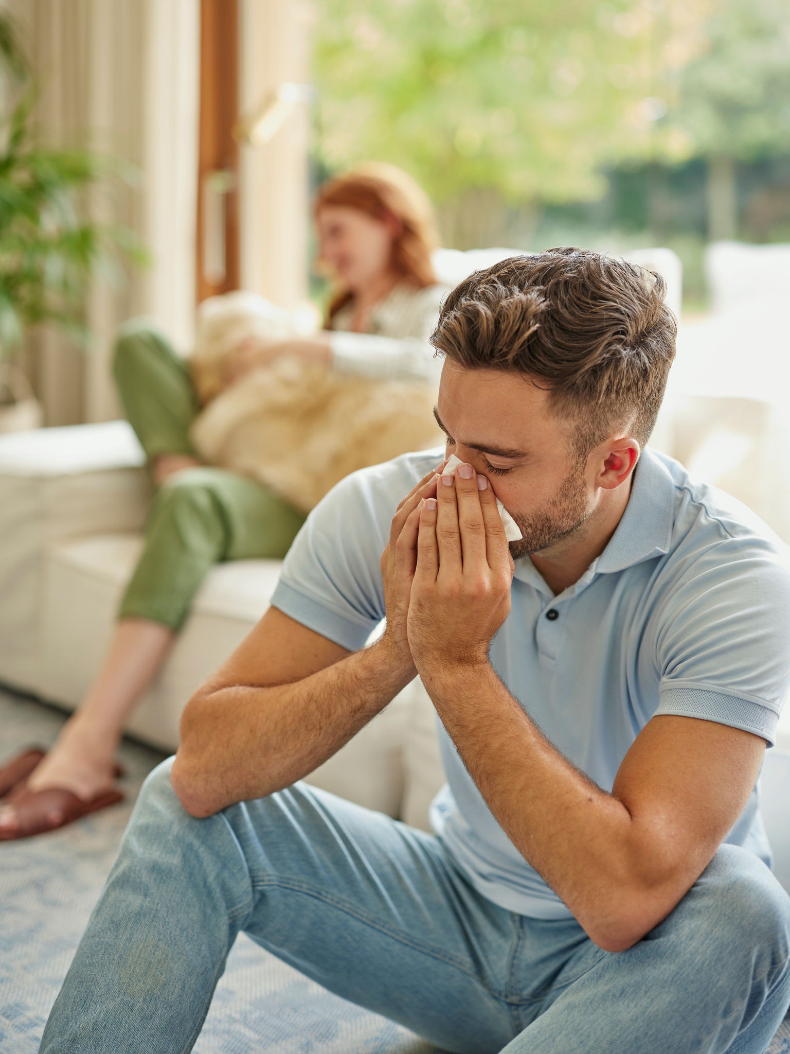 Kvinna i bakgrunden bland textilier med en golden retriever i knät, man i förgrunden som nyser