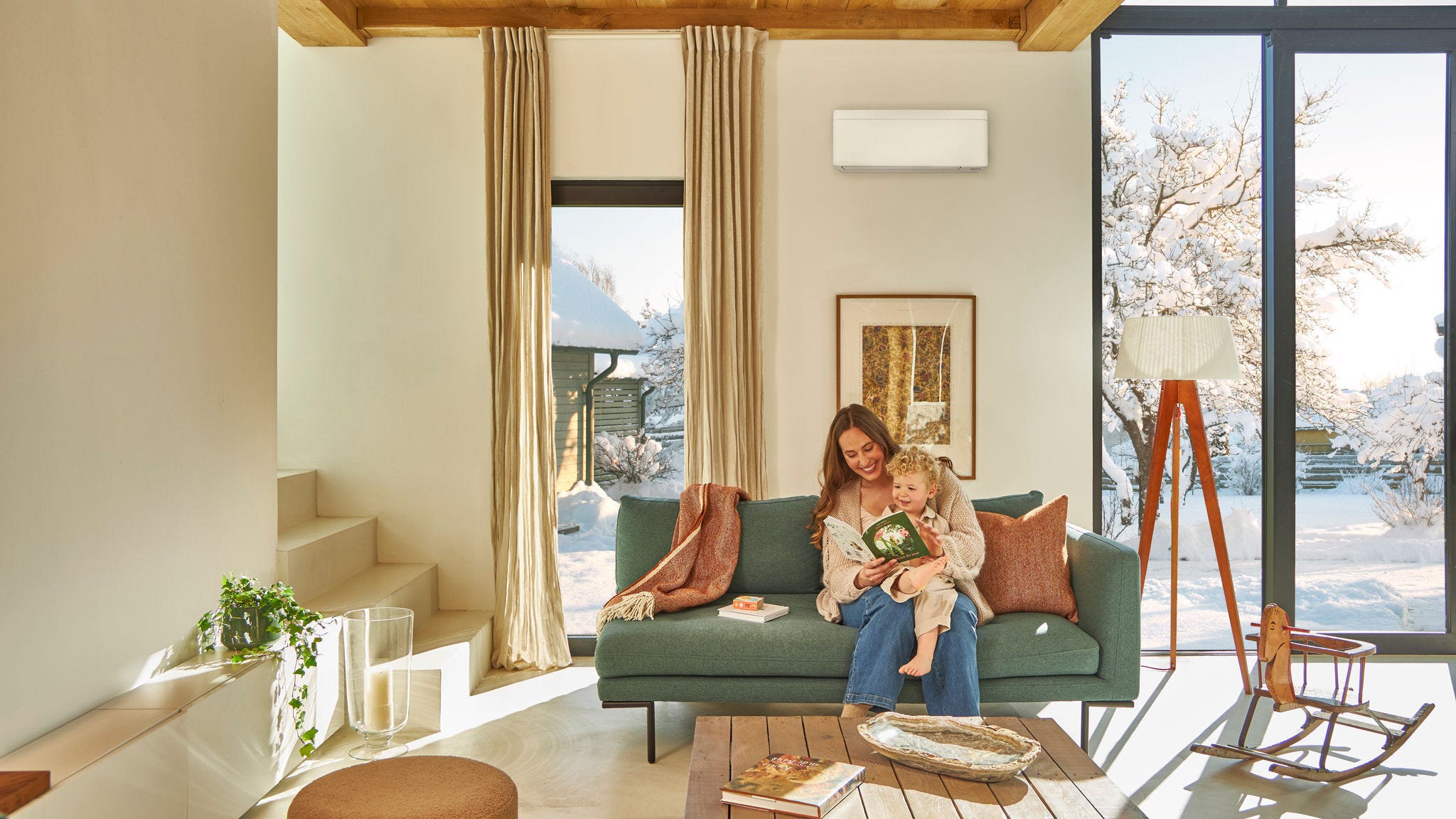 En mamma som sitter på soffan under en väggmonterad luft/luftvärmepump, läser en barnbok med sitt lilla barn i knät, och njuter av värmen medan det snöar utomhus